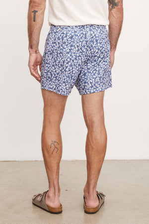 Man in Velvet by Graham & Spencer's RICARDO SWIM SHORT and brown flip-flops, standing with his back to the camera, displaying a tattoo on his right calf.
