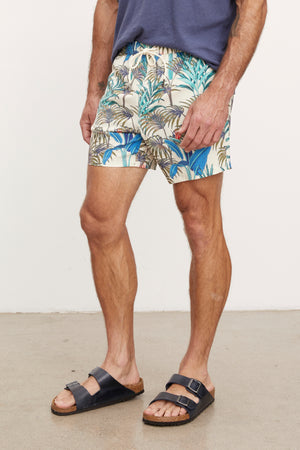 A man standing, wearing colorful floral print RICARDO SWIM SHORTS by Velvet by Graham & Spencer and black slide sandals, showing his legs and part of his torso.