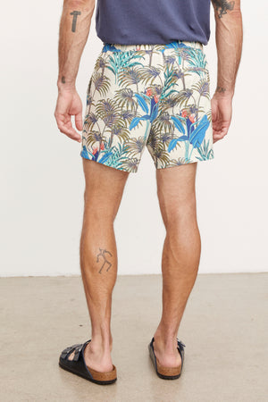 A man stands facing away, wearing RICARDO SWIM SHORT by Velvet by Graham & Spencer and black sandals, showcasing a tattoo on his left calf.