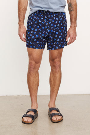 A man wearing Velvet by Graham & Spencer's RICARDO SWIM SHORT swim shorts and black sandals standing against a plain wall. Visible tattoos on his legs.