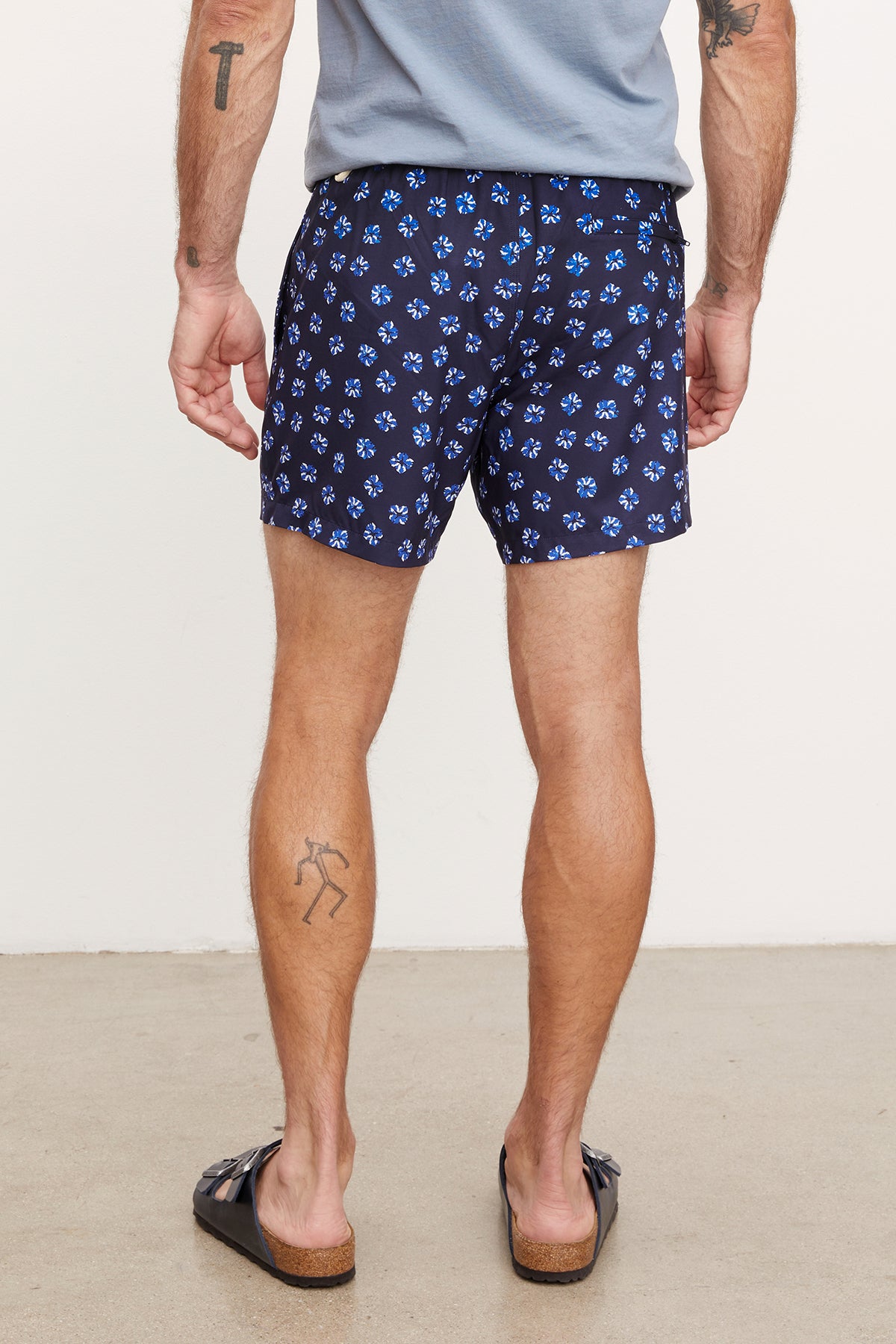   Man wearing Velvet by Graham & Spencer RICARDO SWIM SHORT swim shorts and black flip-flops, standing and showing the side and rear view, with visible tattoos on both arms and left leg. 