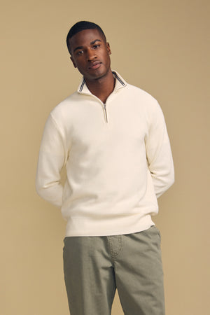 A man stands in front of a beige background, wearing the Velvet by Graham & Spencer BRENTWOOD SWEATER—a cream quarter-zip style—paired with light olive pants, his hands behind his back as he looks at the camera.