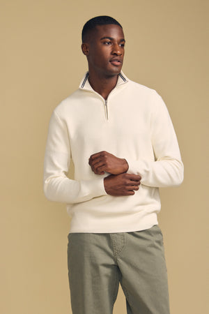 A man stands against a beige background, wearing the BRENTWOOD SWEATER by Velvet by Graham & Spencer—a cream-colored quarter-zip—paired with light olive pants. He looks slightly to the side, hands lightly clasped.