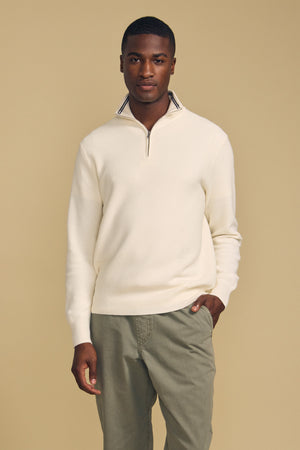 A man stands against a beige background, wearing the BRENTWOOD SWEATER by Velvet by Graham & Spencer—a cream quarter-zip style—paired with light olive pants and one hand in his pocket.