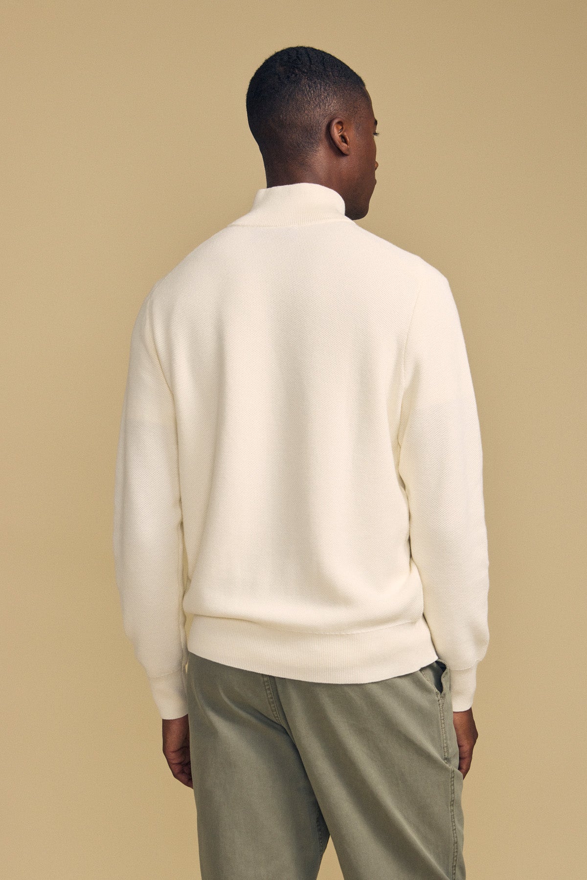 A person stands facing away against a beige background, wearing the cream-colored BRENTWOOD SWEATER by Velvet by Graham & Spencer and olive green pants.
