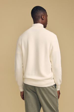 A person stands facing away against a beige background, wearing the cream-colored BRENTWOOD SWEATER by Velvet by Graham & Spencer and olive green pants.