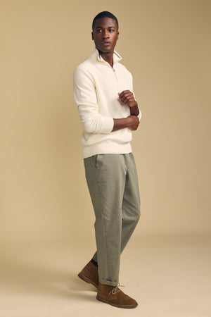 A person stands against a beige background, wearing the Velvet by Graham & Spencer BRENTWOOD SWEATER in cream, paired with light green trousers and brown suede shoes, with one arm crossed and the other touching their collar.