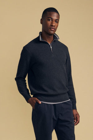 A man stands against a beige background, wearing the Velvet by Graham & Spencer BRENTWOOD SWEATER—a dark quarter-zip style—layered over a light undershirt and paired with dark pants, with one hand in his pocket.