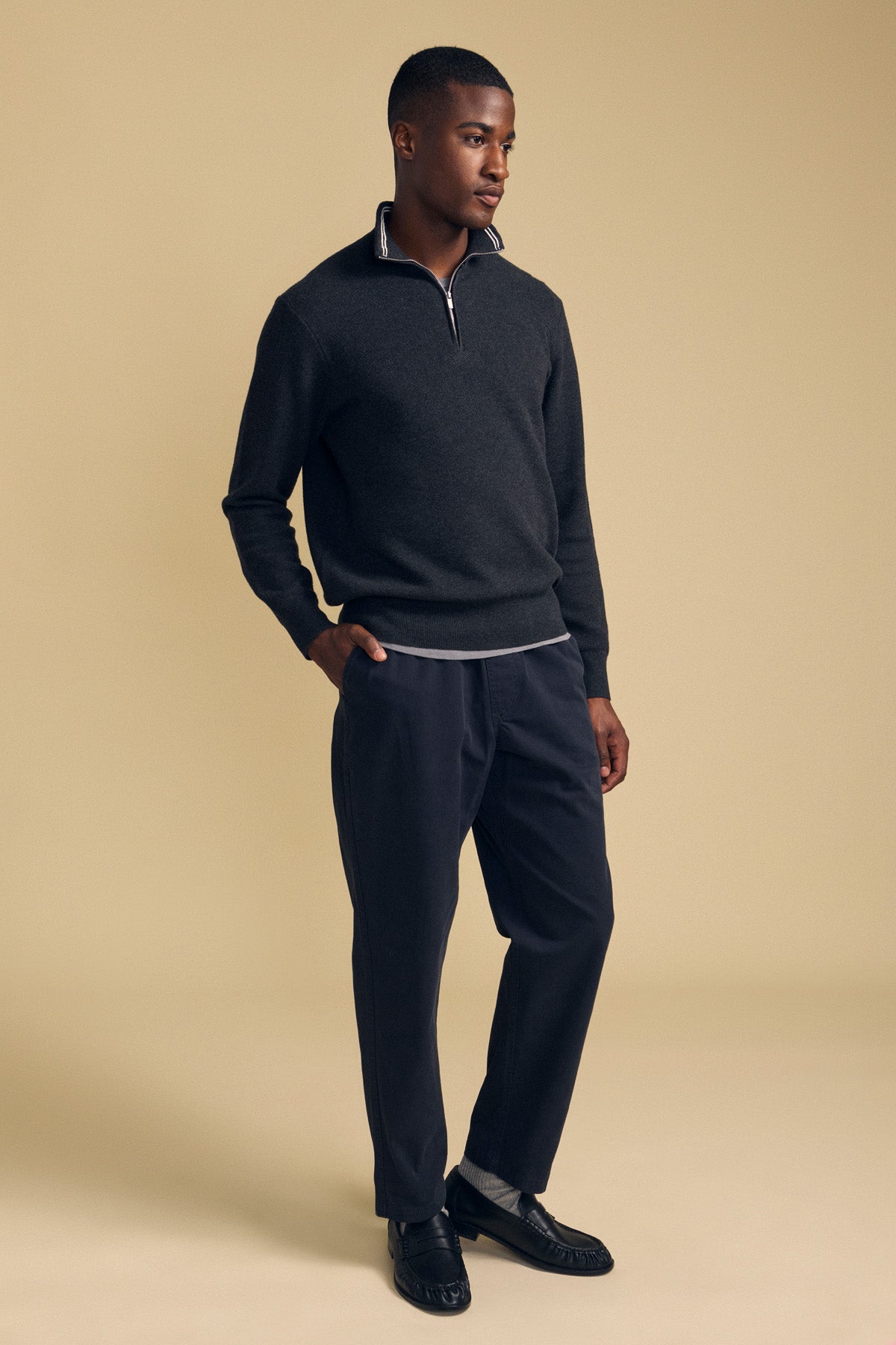 A man stands against a beige background wearing the BRENTWOOD SWEATER by Velvet by Graham & Spencer, paired with dark trousers and black loafers, with his left hand in his pocket.