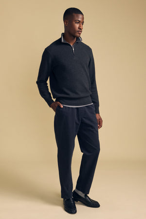 A man stands against a beige background wearing the BRENTWOOD SWEATER by Velvet by Graham & Spencer, paired with dark trousers and black loafers, with his left hand in his pocket.