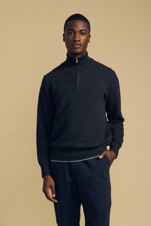 A person stands against a plain beige background, wearing the BRENTWOOD SWEATER by Velvet by Graham & Spencer— a dark quarter-zip style—paired with dark pants and one hand in their pocket.