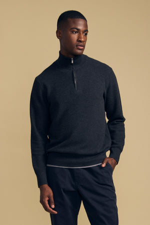 A man wearing the Velvet by Graham & Spencer BRENTWOOD SWEATER and dark pants stands against a plain beige background with one hand in his pocket.