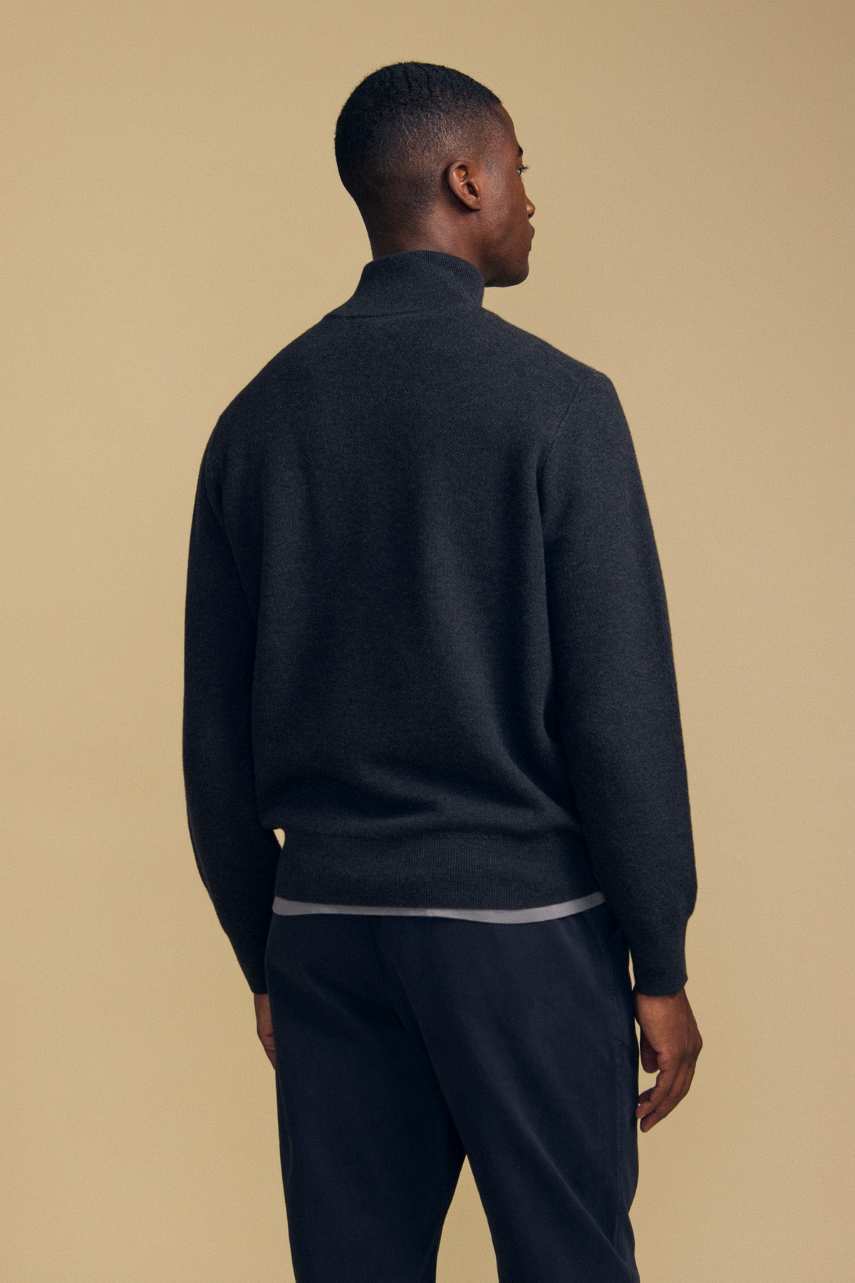 A man stands facing away against a plain beige background, wearing the Velvet by Graham & Spencer BRENTWOOD SWEATER in dark gray with long sleeves, paired with dark pants.-41859621290177