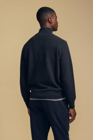 A man stands facing away against a plain beige background, wearing the Velvet by Graham & Spencer BRENTWOOD SWEATER in dark gray with long sleeves, paired with dark pants.