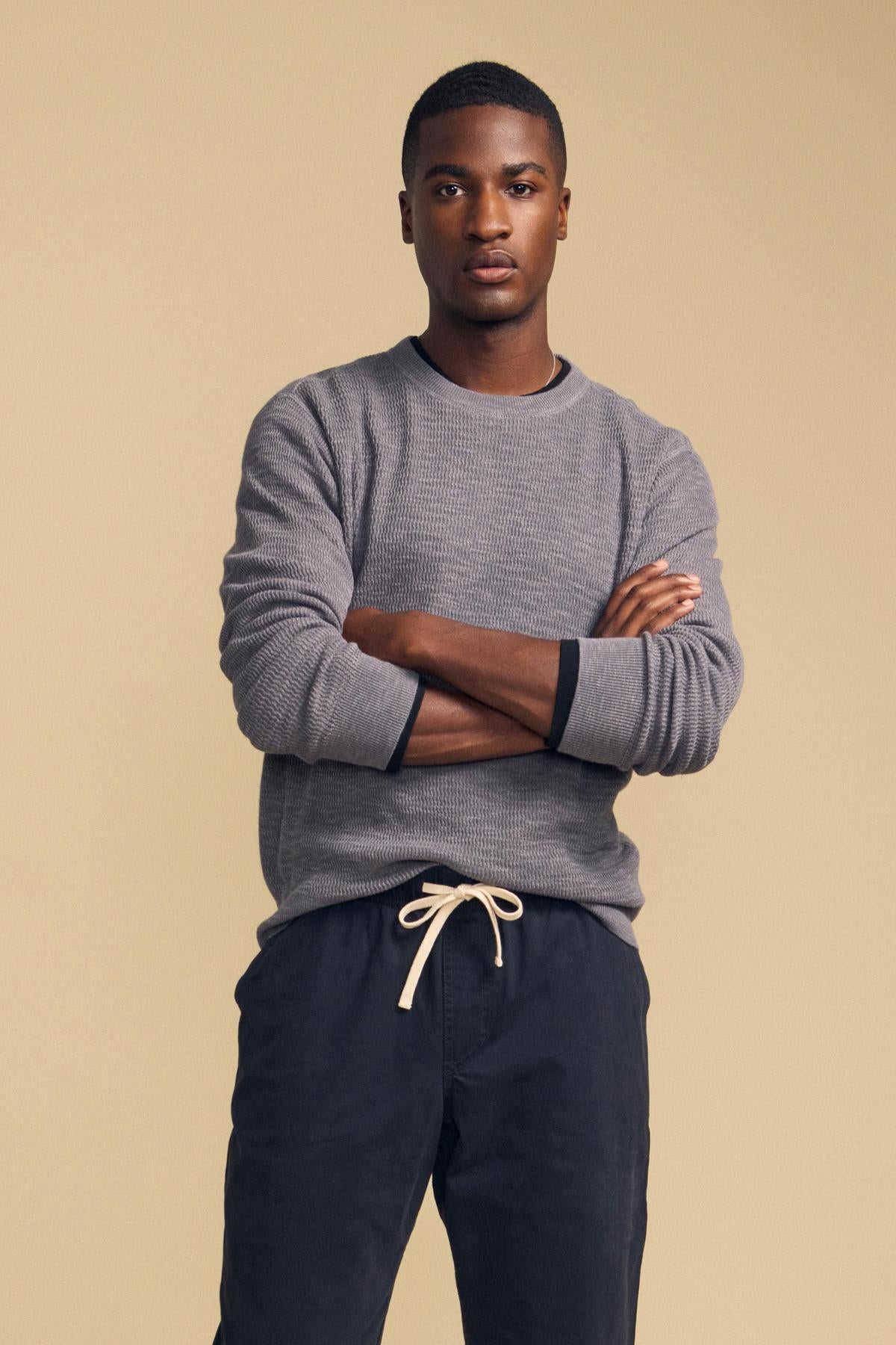 A man wearing a grey CONOR SWEATER with dark cuffs and navy blue drawstring pants, standing with his arms crossed.-46287883272385