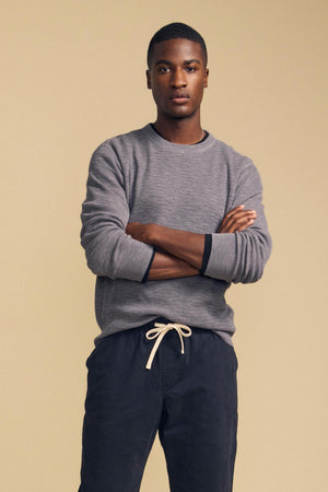 A man wearing a grey CONOR SWEATER with dark cuffs and navy blue drawstring pants, standing with his arms crossed.