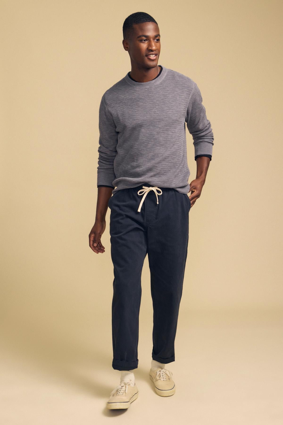A man wearing a grey long-sleeved textured CONOR SWEATER by Velvet by Graham & Spencer, dark drawstring pants, and canvas sneakers.