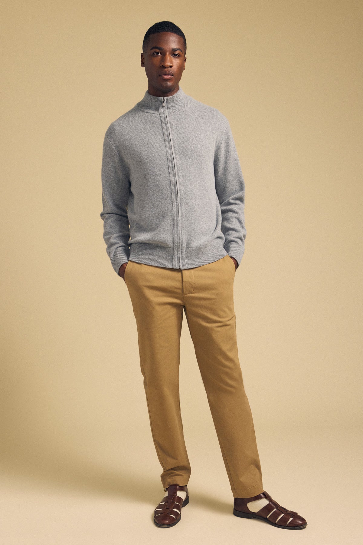 A man wearing a grey DALLEN FULL-ZIP SWEATER, tan trousers, and brown leather sandals with his hands in his pockets.