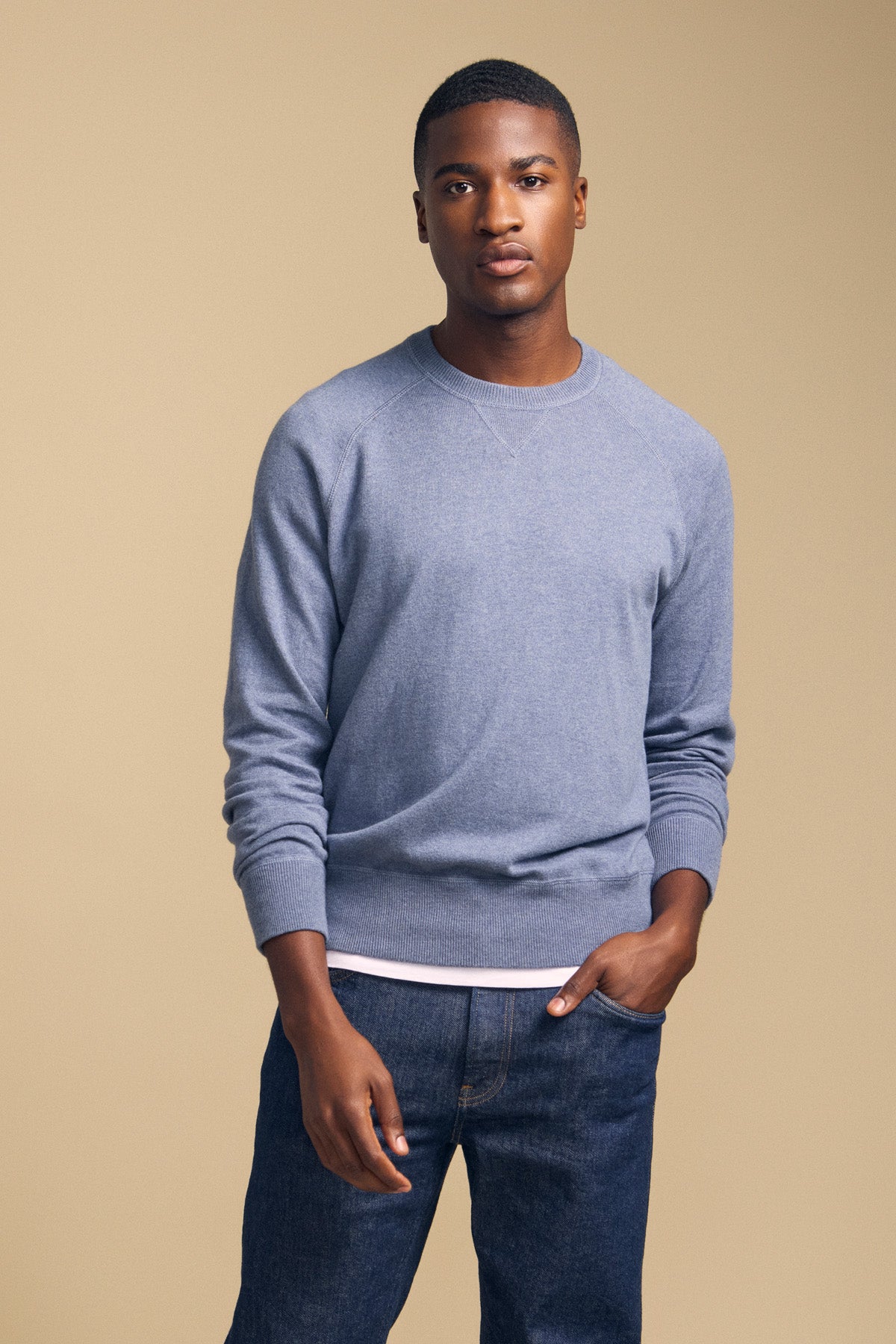 A person stands against a beige background, wearing the DARIO SWEATER by Velvet by Graham & Spencer—a light blue cotton-cashmere crewneck—layered over a white shirt and paired with dark jeans, one hand in their pocket.