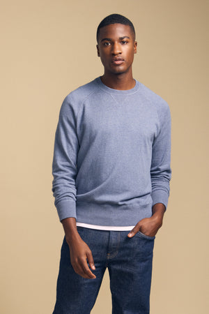 A person stands against a beige background, wearing the DARIO SWEATER by Velvet by Graham & Spencer—a light blue cotton-cashmere crewneck—layered over a white shirt and paired with dark jeans, one hand in their pocket.