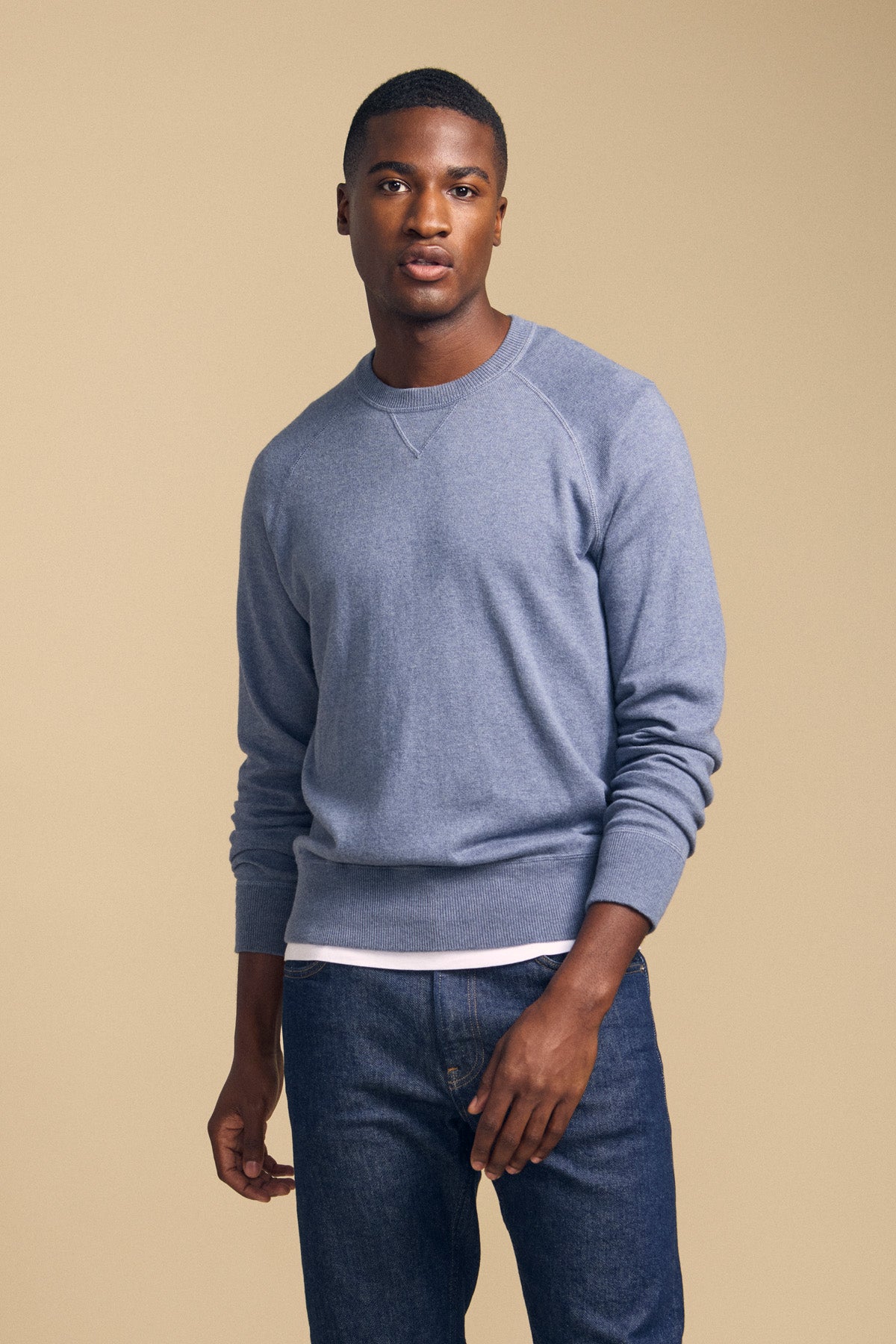A man stands against a beige background, wearing the Velvet by Graham & Spencer DARIO SWEATER—a light blue, mid-weight crewneck—layered over a white shirt and paired with blue jeans.-42092466012353