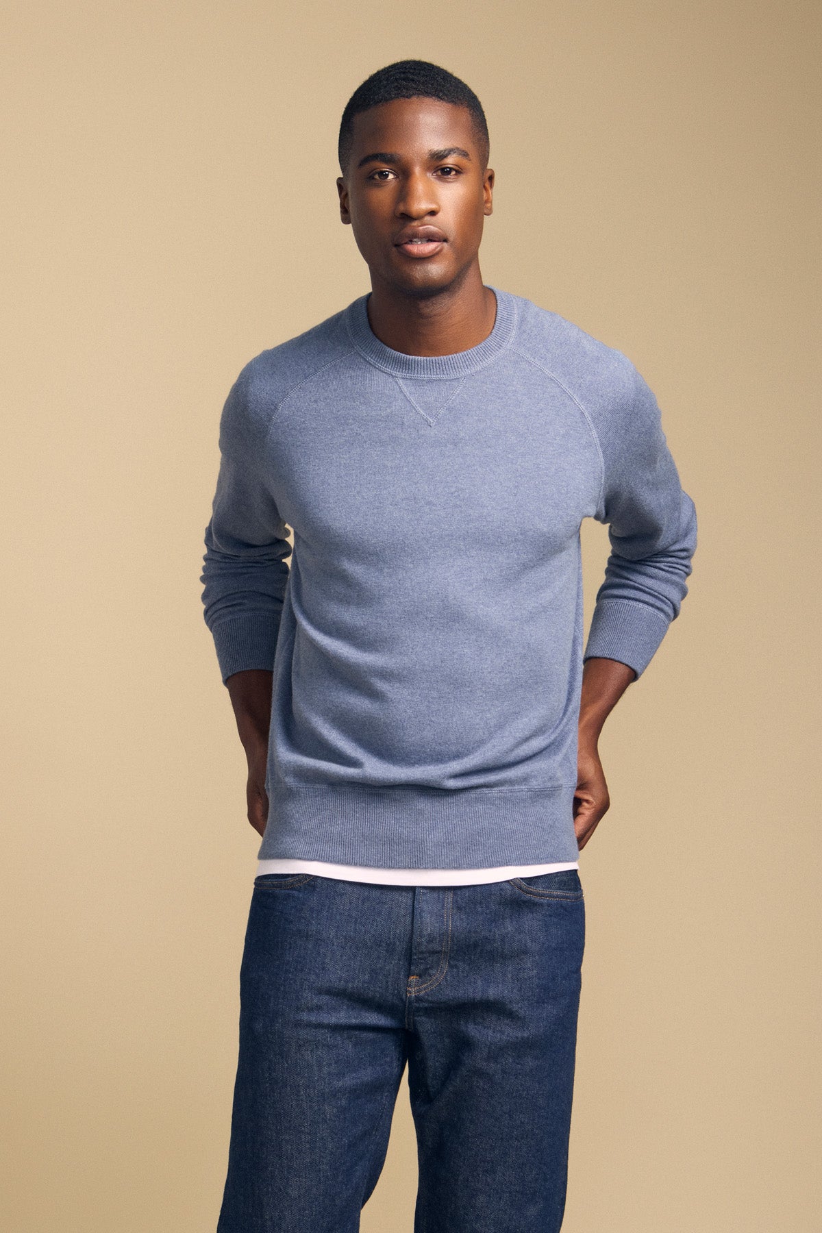 A man stands against a plain beige background, wearing the DARIO SWEATER by Velvet by Graham & Spencer—a blue, crew-neckline mid-weight sweater—layered over a white shirt and paired with blue jeans, with his hands behind his back.
