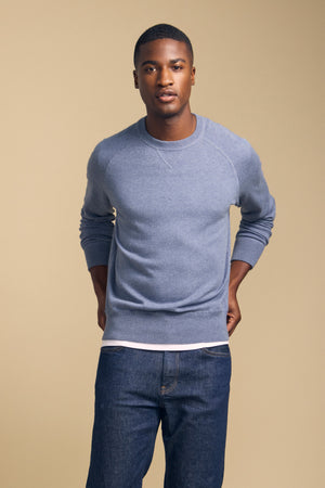 A man stands against a plain beige background, wearing the DARIO SWEATER by Velvet by Graham & Spencer—a blue, crew-neckline mid-weight sweater—layered over a white shirt and paired with blue jeans, with his hands behind his back.