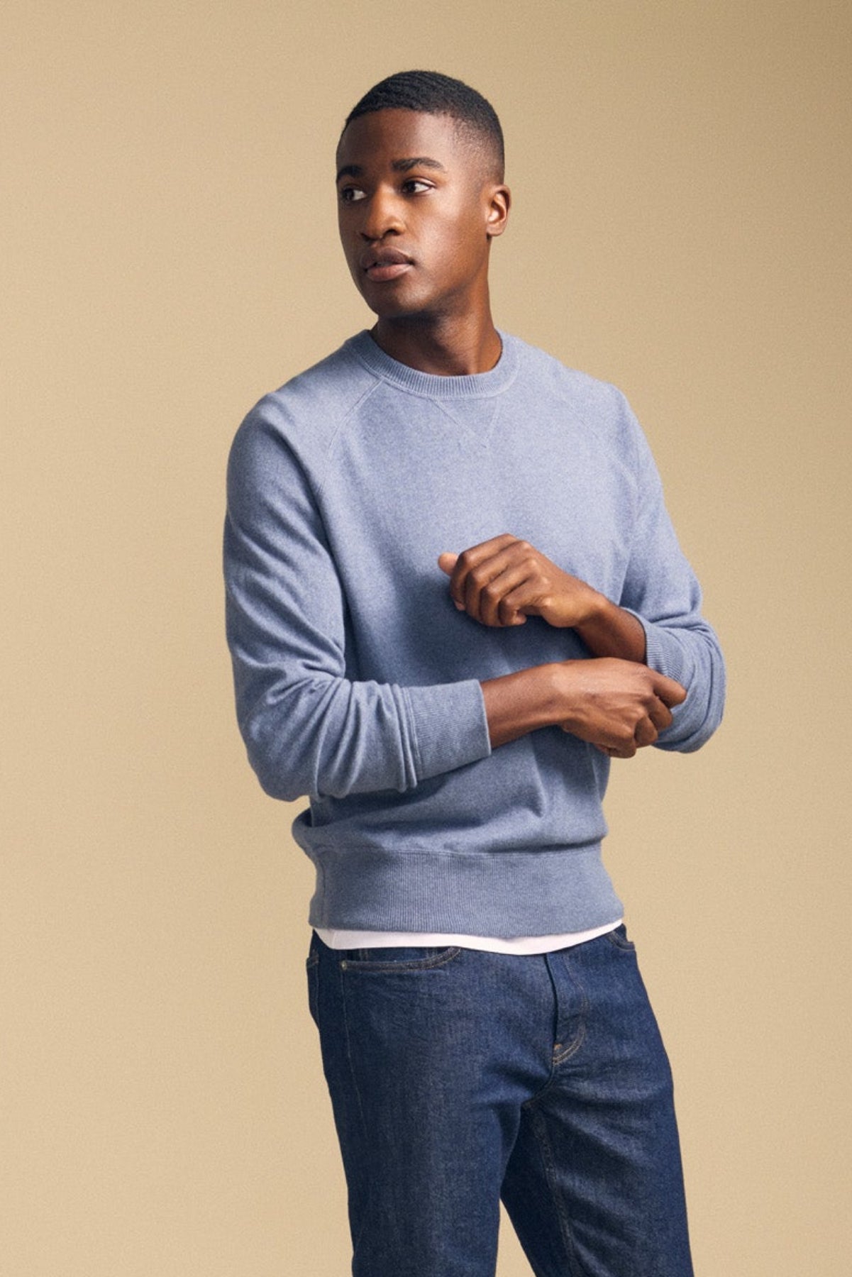 A young man stands against a beige background, looking to his left as he adjusts his sleeve, wearing the DARIO SWEATER—a blue, mid-weight crewneck by Velvet by Graham & Spencer—paired with jeans.