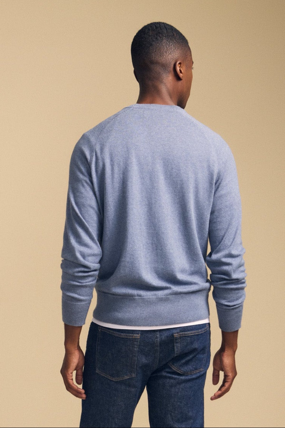 A man stands facing away from the camera, wearing the Velvet by Graham & Spencer DARIO SWEATER—a light blue mid-weight cotton cashmere pullover featuring a classic crew neckline—paired with dark jeans, set against a plain beige background.