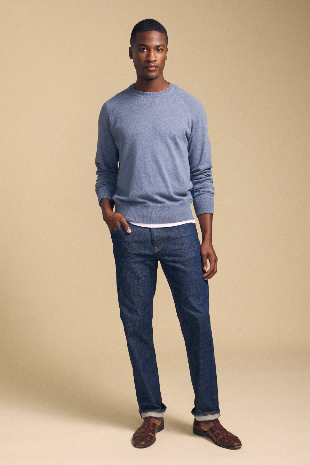 A man stands against a beige background, wearing the DARIO SWEATER from Velvet by Graham & Spencer—a light blue cotton cashmere knit with a classic crew neckline—layered over a white shirt, paired with blue jeans and brown sandals.-42092465750209