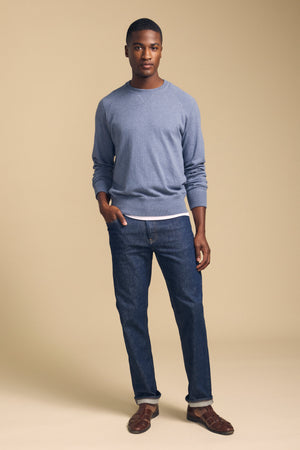 A man stands against a beige background, wearing the DARIO SWEATER from Velvet by Graham & Spencer—a light blue cotton cashmere knit with a classic crew neckline—layered over a white shirt, paired with blue jeans and brown sandals.