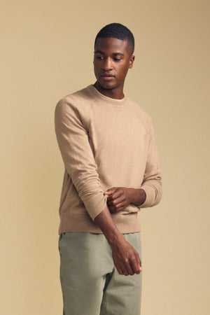 A man wearing the DARIO SWEATER by Velvet by Graham & Spencer—a beige, mid-weight crew neck sweater—paired with light green pants stands against a plain beige background, adjusting his sleeve with his left hand while looking slightly to his right.