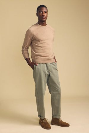 A man stands against a beige background wearing the Velvet by Graham & Spencer DARIO SWEATER—a tan cotton cashmere style with a crew neckline—paired with light olive pants and brown suede shoes, his hands in his pockets.