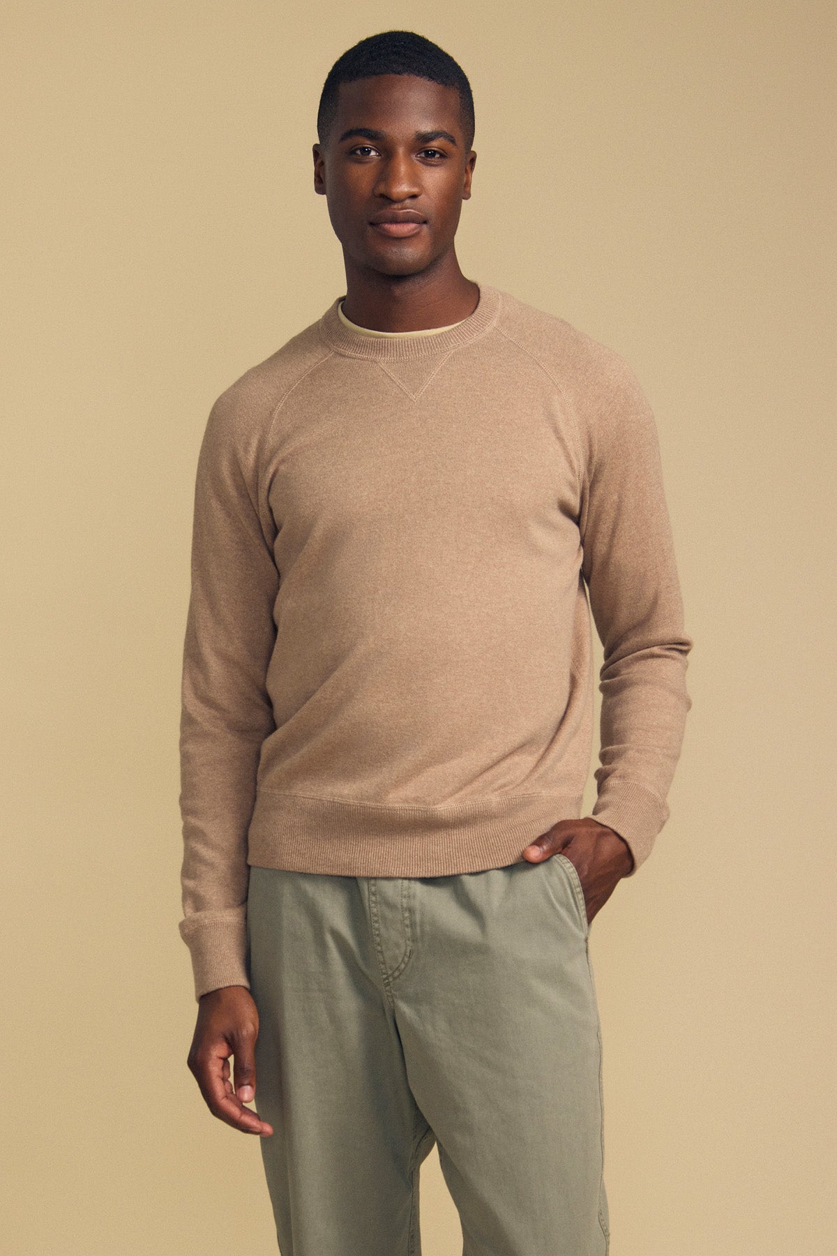 A person stands against a beige background wearing the DARIO SWEATER by Velvet by Graham & Spencer—a light brown cotton-cashmere blend with a classic crew neckline—paired with light gray pants and one hand in their pocket.