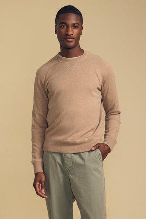 A person stands against a beige background wearing the DARIO SWEATER by Velvet by Graham & Spencer—a light brown cotton-cashmere blend with a classic crew neckline—paired with light gray pants and one hand in their pocket.