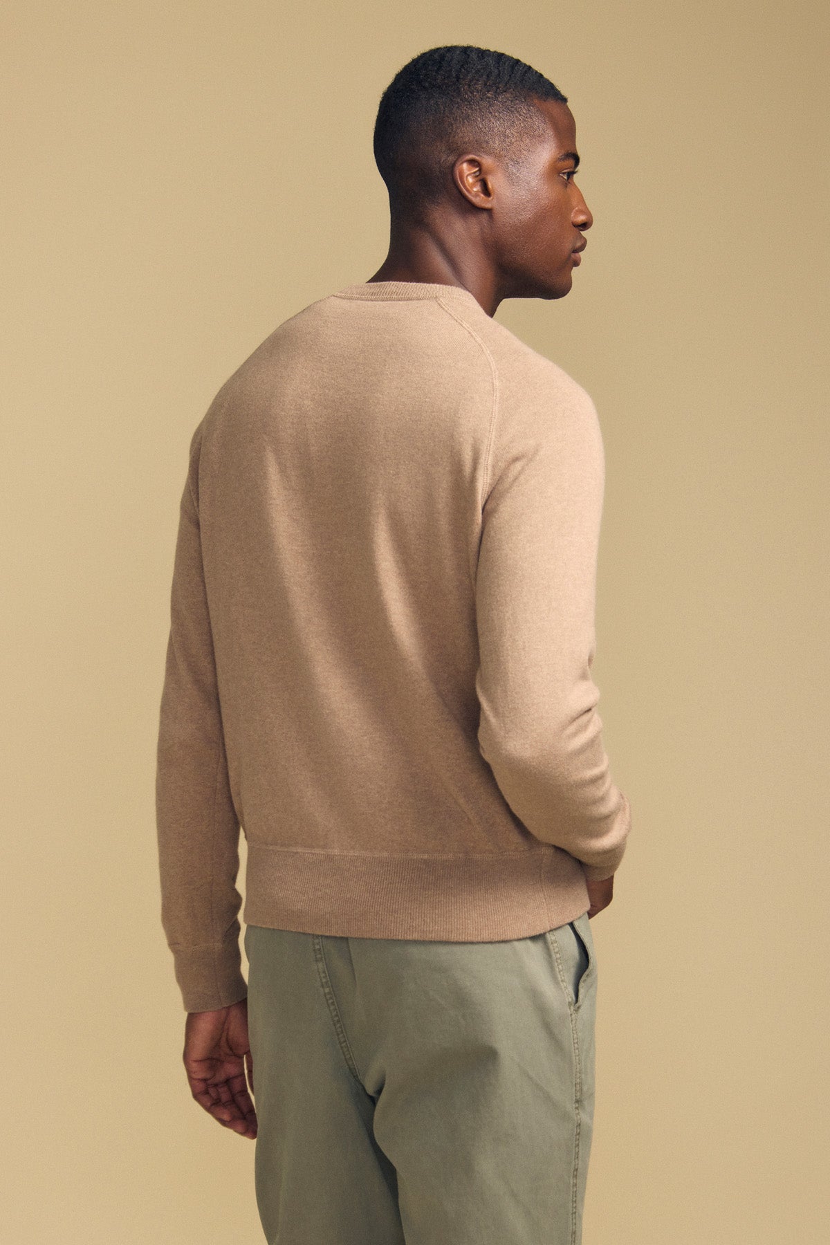 A man stands facing away against a plain beige background, wearing the DARIO SWEATER by Velvet by Graham & Spencer—a tan, crew-neckline mid-weight sweater—paired with light green pants.
