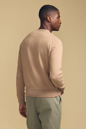 A man stands facing away against a plain beige background, wearing the DARIO SWEATER by Velvet by Graham & Spencer—a tan, crew-neckline mid-weight sweater—paired with light green pants.