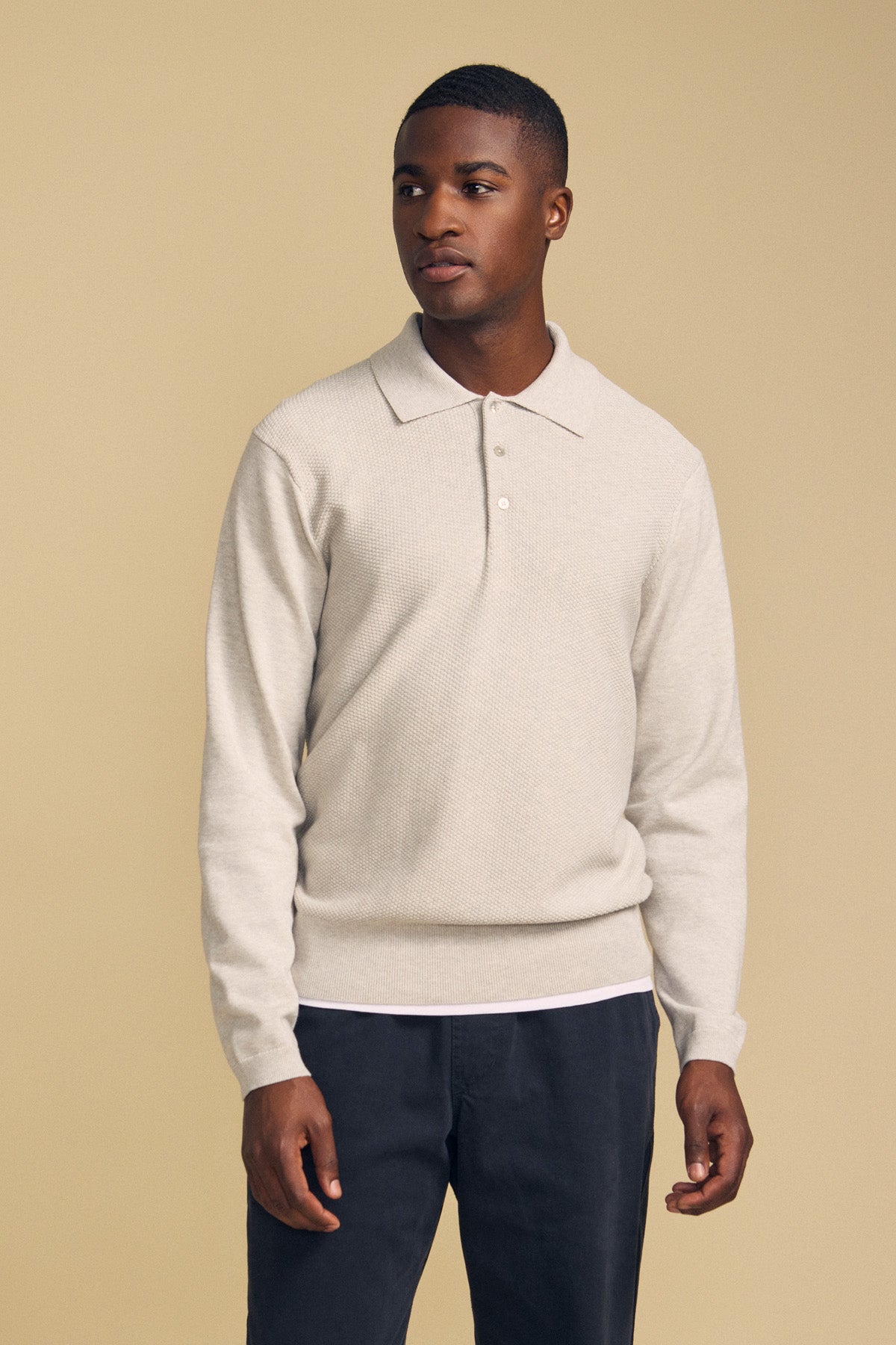 A man wearing the KINGSLEY SWEATER POLO by Velvet by Graham & Spencer in light beige with dark pants stands against a plain beige background, looking off to the side—this refined men’s layering piece is perfect for any occasion.