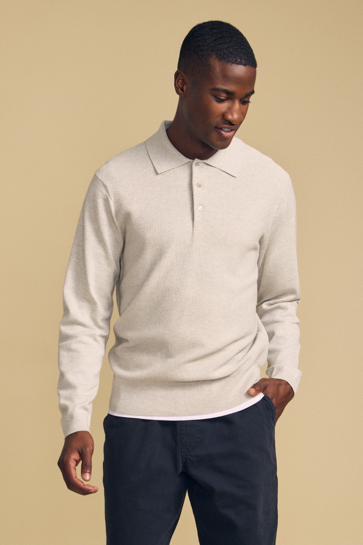 A man wearing the KINGSLEY SWEATER POLO by Velvet by Graham & Spencer—a light beige cotton-cashmere sweater polo featuring a refined knit collar—pairs it with dark pants and stands against a plain beige background, looking down with a slight smile, effortlessly highlighting this versatile men’s layering piece.-42092016730305