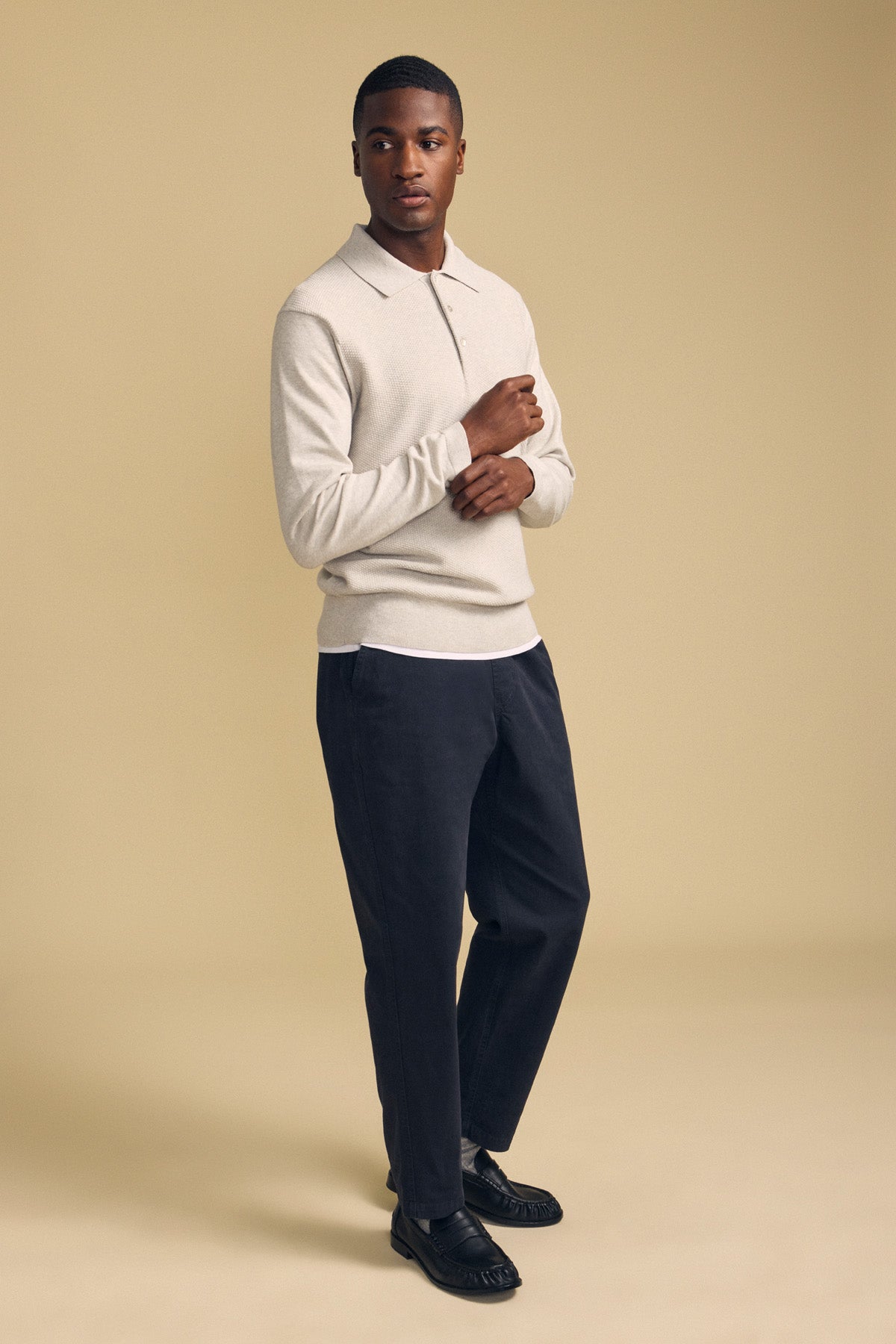 A man stands against a beige background wearing the KINGSLEY SWEATER POLO by Velvet by Graham & Spencer—a light beige cotton-cashmere top with a refined knit collar—paired with dark trousers and black shoes, making it the perfect men’s layering piece for effortless style.