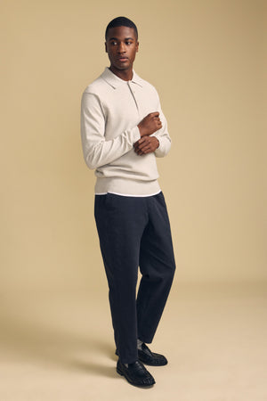 A man stands against a beige background wearing the KINGSLEY SWEATER POLO by Velvet by Graham & Spencer—a light beige cotton-cashmere top with a refined knit collar—paired with dark trousers and black shoes, making it the perfect men’s layering piece for effortless style.