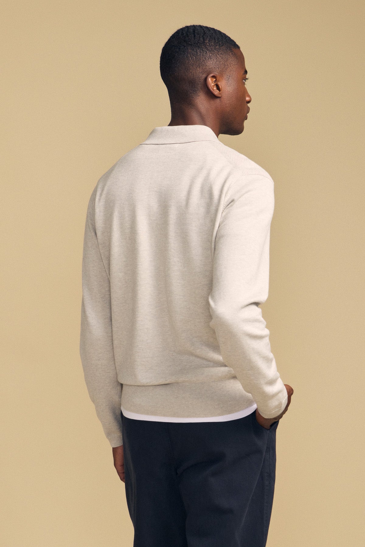 A person stands facing away from the camera wearing the light beige KINGSLEY SWEATER POLO by Velvet by Graham & Spencer and dark pants against a plain beige background.-42092016828609