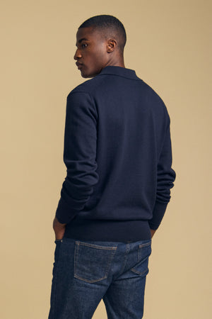 A man stands with his back turned, wearing the KINGSLEY SWEATER POLO in dark navy from Velvet by Graham & Spencer and blue jeans against a plain beige background—the perfect men’s layering piece for effortless, everyday style.