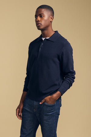 A man stands against a beige background, wearing the Velvet by Graham & Spencer KINGSLEY SWEATER POLO in dark navy—a cotton-cashmere piece featuring a knit collar—paired with blue jeans. With one hand in his pocket and a neutral expression, he showcases this versatile men’s layering essential that blends comfort and style effortlessly.