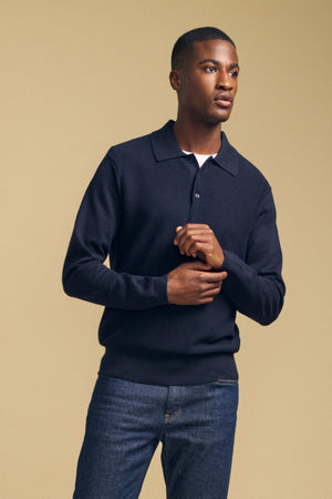 A person stands against a beige background, wearing the KINGSLEY SWEATER POLO by Velvet by Graham & Spencer—a dark cotton-cashmere sweater with a knit collar—paired with blue jeans, looking slightly to the side with hands loosely clasped; this refined men’s layering piece is perfect for any occasion.