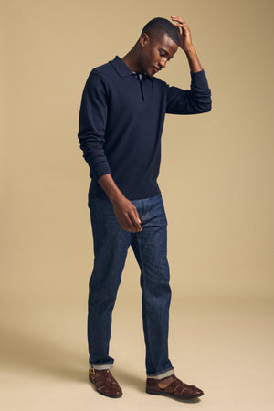 A man wearing the KINGSLEY SWEATER POLO by Velvet by Graham & Spencer—a navy cotton-cashmere knit with a refined collar—pairs it with dark blue jeans and brown sandals as he stands against a beige background, touching his head and looking down. This versatile men’s layering piece elevates his relaxed outfit with its sophisticated style.