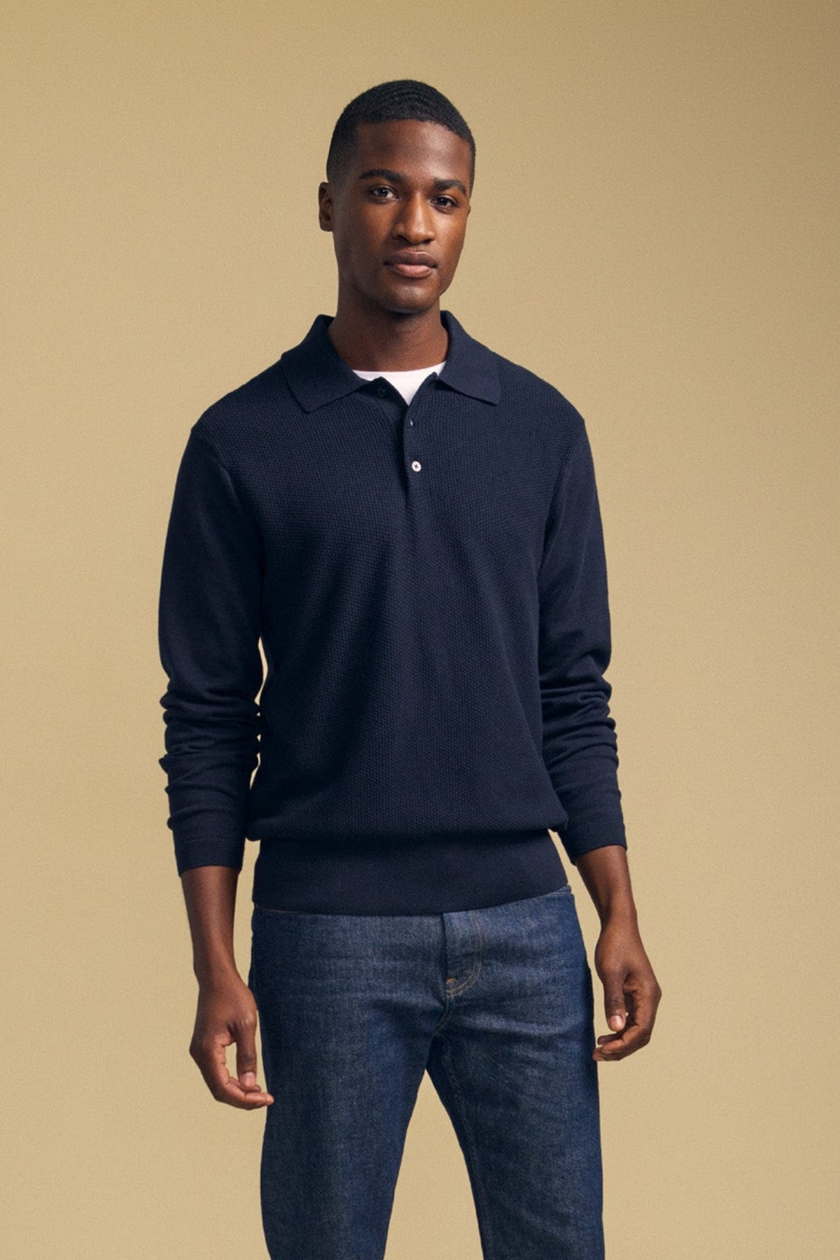 A man stands against a plain beige background, wearing the KINGSLEY SWEATER POLO by Velvet by Graham & Spencer over a white undershirt and blue jeans—a versatile men’s layering piece perfect for effortless style.-42092017057985