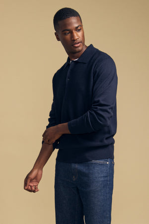 A man wearing the KINGSLEY SWEATER POLO by Velvet by Graham & Spencer—a navy cotton-cashmere blend with a refined knit collar—pairs it with blue jeans as he stands against a plain beige background, looking to his left and adjusting his sleeve, showcasing this versatile men’s layering piece.