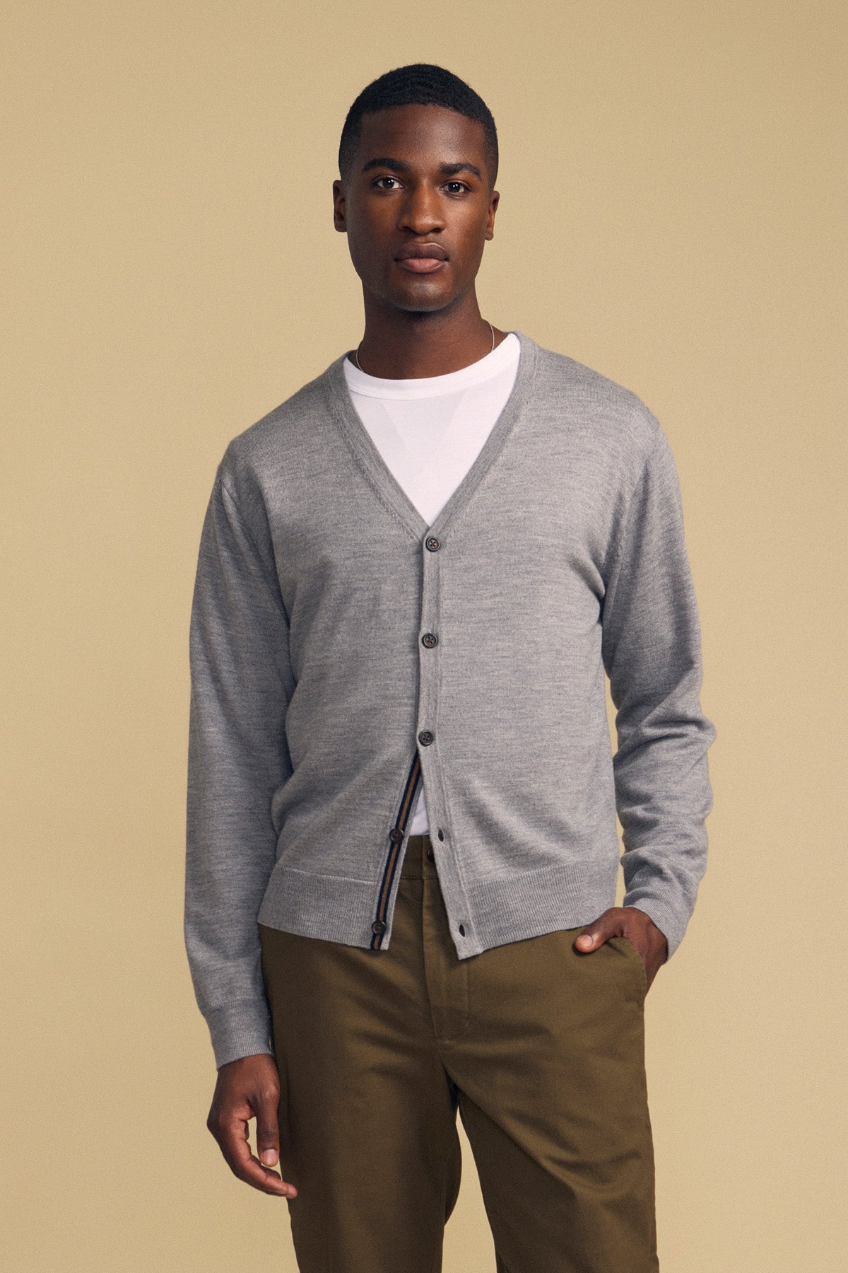 A man stands against a beige background wearing the SILVIO CARDIGAN by Velvet by Graham & Spencer—a gray V-neck cardigan with ribbed trim—layered over a white t-shirt and paired with olive green pants.-42092513231041