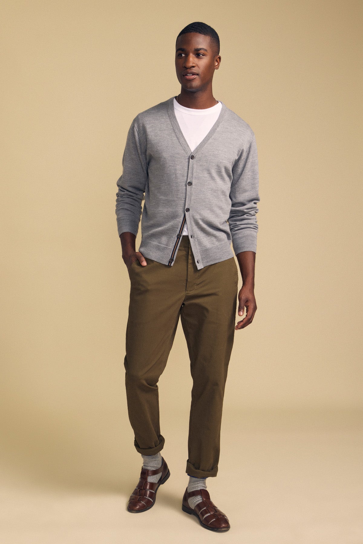 A man stands against a beige background wearing the SILVIO CARDIGAN by Velvet by Graham & Spencer over a white t-shirt, paired with brown pants, gray socks, and brown sandals.